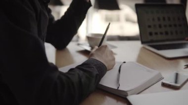 Kafenin ahşap masasında oturan, elinde kalem ve defter olan bir adamın elleri..