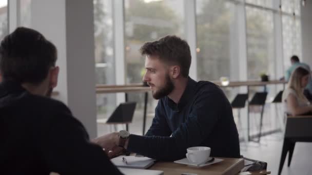 Dos hombres de negocios alegres discutiendo algo y sonriendo mientras se sienta en la cafetería con un ordenador portátil y teléfonos. Dos jóvenes colegas exitosos discutiendo momentos de organización por mesa en la cafetería . — Vídeos de Stock