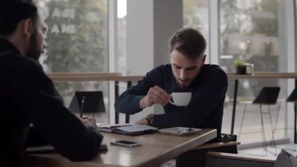 Zwei gut gelaunte Geschäftsleute diskutieren etwas und lächeln, während sie mit Laptop und Handy im Café sitzen. zwei junge erfolgreiche Kollegen im Gespräch über Organisation Momente von Tisch zu Tisch im Café. — Stockvideo