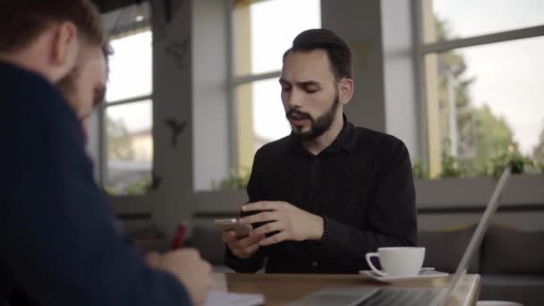 Deux hommes d'affaires joyeux discutant de quelque chose et souriant tout en étant assis au café avec un ordinateur portable et des téléphones. Deux jeunes collègues qui ont réussi à discuter des moments d'organisation par table dans un café . — Video