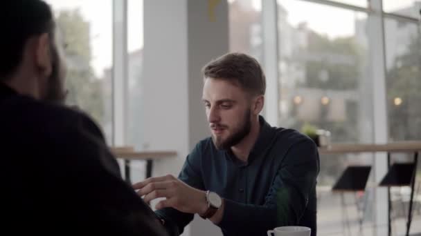 Två glada affärsman diskuterar något och ler medan du sitter på caféet med en bärbar dator och telefoner. Två unga framgångsrika kollegor diskuterar organisationsmoment vid bord på caféet. — Stockvideo