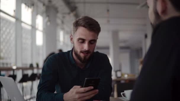 Två glada affärsman diskuterar något och ler medan du sitter på caféet med en bärbar dator och telefoner. Två unga framgångsrika kollegor diskuterar organisationsmoment vid bord på caféet. — Stockvideo