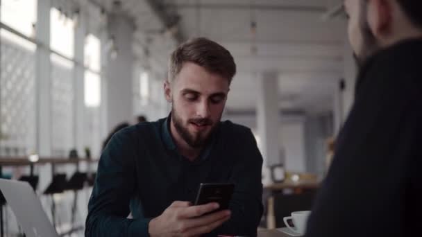 Deux hommes d'affaires joyeux discutant de quelque chose et souriant tout en étant assis au café avec un ordinateur portable et des téléphones. Deux jeunes collègues qui ont réussi à discuter des moments d'organisation par table dans un café . — Video