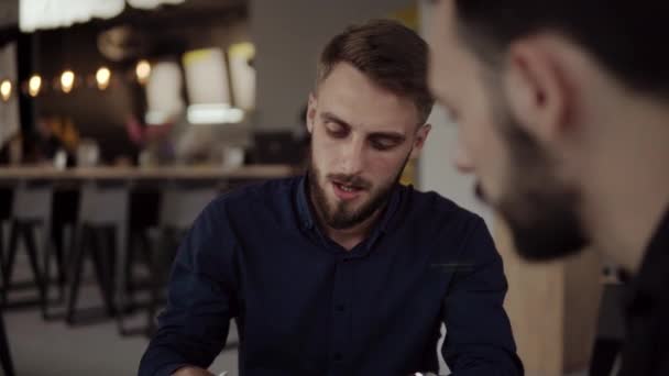 Két vidám üzletember megvitat valamit és mosolyog, miközben a kávézóban ülnek egy laptoppal és telefonnal. Két fiatal, sikeres kolléga a szervezési pillanatok asztali megvitatásában a kávézóban. — Stock videók
