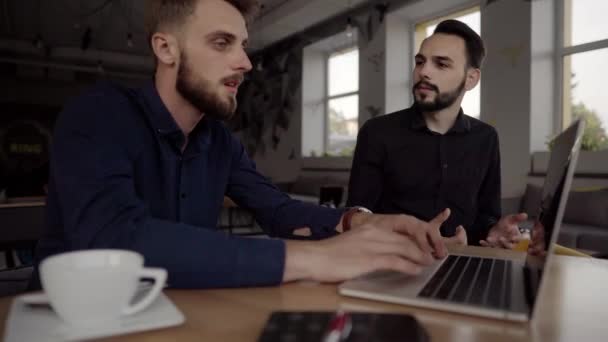 Zwei gut gelaunte Geschäftsleute diskutieren etwas und lächeln, während sie mit Laptop und Handy im Café sitzen. zwei junge erfolgreiche Kollegen im Gespräch über Organisation Momente von Tisch zu Tisch im Café. — Stockvideo