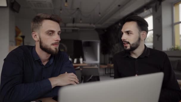 Dois homens de negócios alegres discutindo algo e sorrindo enquanto estavam sentados no café com um laptop e telefones. Dois jovens colegas de sucesso em discutir momentos de organização por mesa no café . — Vídeo de Stock