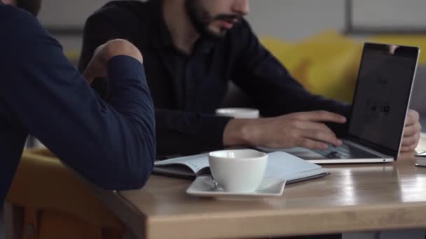 Dos hombres de negocios alegres discutiendo algo y sonriendo mientras se sienta en la cafetería con un ordenador portátil y teléfonos. Dos jóvenes colegas exitosos discutiendo momentos de organización por mesa en la cafetería . — Vídeos de Stock