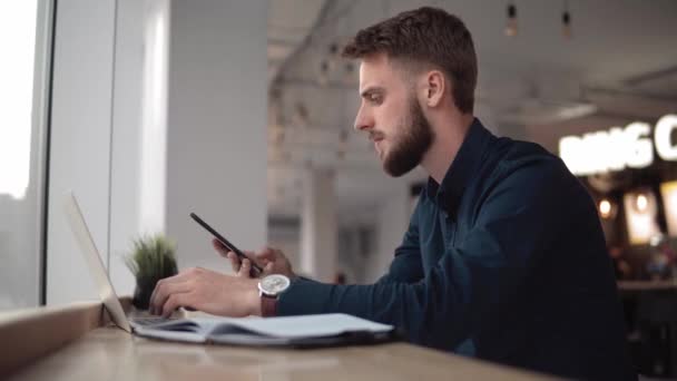 Mladý pohledný podnikatel sedící před svým zápisníkem v útulku a začít používat smartphone pro rozhovor se svým obchodním partnerem nebo kolegy. — Stock video