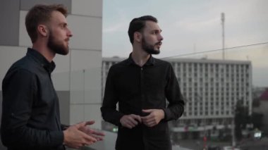 Genç iş arkadaşları gülerek bir şeyler tartışıyor ve akşamları yüksek bir yerde, gün batımında dışarıda dikiliyorlar. İş ortaklığı ya da iş arkadaşı diyaloğu.