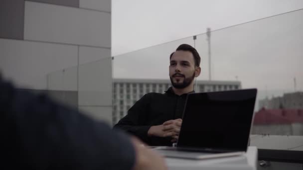 Two business mans discussing something and smiling while sitting outdoor with a laptop and phones in the evening time. Two young successful colleagues in discussing organizational moments. — Stock Video