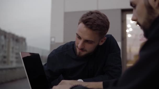 Dos hombres de negocios discutiendo algo y sonriendo mientras se sienta al aire libre con un ordenador portátil y teléfonos en el tiempo de la noche. Dos jóvenes colegas exitosos discutiendo momentos organizacionales . — Vídeos de Stock