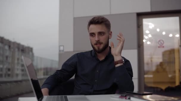 Dos hombres de negocios discutiendo algo y sonriendo mientras se sienta al aire libre con un ordenador portátil y teléfonos en el tiempo de la noche. Dos jóvenes colegas exitosos discutiendo momentos organizacionales . — Vídeo de stock