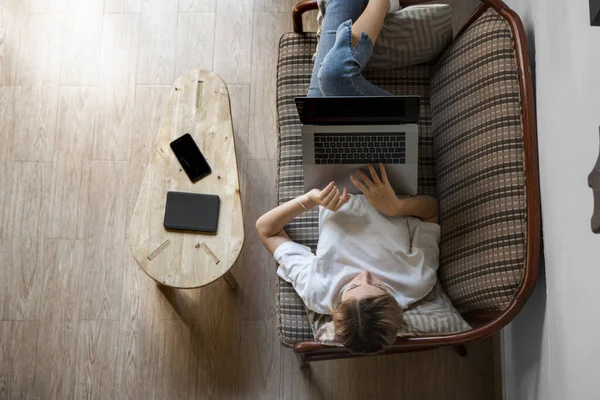 Mujer con un portátil acostada en un sofá. Estudiar y trabajar en línea, freelance. Mujer autónoma, niña que trabaja con su cuaderno acostada en un sofá con un teléfono, smartphone y lector de libros en la mesa . — Foto de Stock