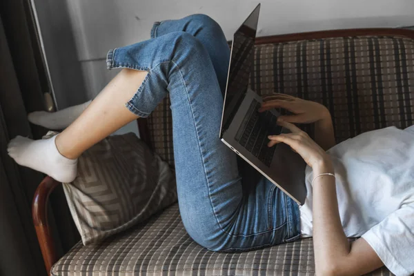 Woman with a laptop laying on a sofa. Study and work online, freelance. Self employed woman, girl working with her notebook laying on a couch with a phone, smartphone and ereader on table. — Stock Photo, Image