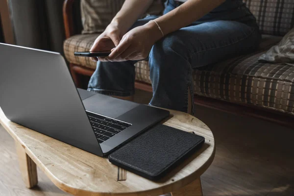 Mujer con un ordenador portátil está sentado en un sofá y utilizar un teléfono inteligente. Estudiar y trabajar en línea, freelance. Chica autónoma está trabajando con su cuaderno sentado en un sofá con un teléfono y leer en la mesa . — Foto de Stock