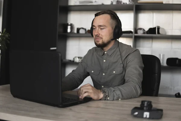 年轻的商人或学生穿着衬衫坐在耳机里，与电脑显示器对一起。在办公室的桌子上用一张电脑，用深思熟虑的表情. — 图库照片