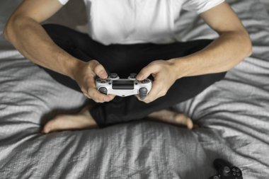 Beyaz tişörtlü bir oyuncu elinde bir av tüfeği tutuyor ve bir kanepede oturuyor. Adam elleri beyaz bir kumandayla video oyunu oynuyor. Evde zevkle bir video oyunu oynayın..