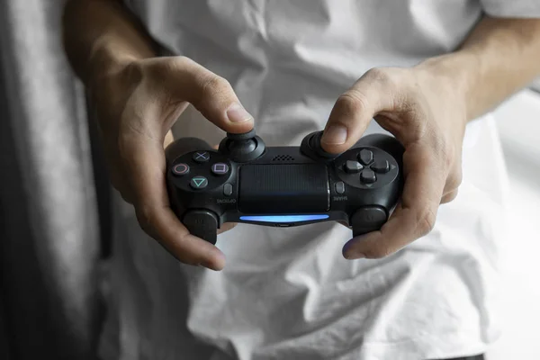 Gamer en camiseta blanca sosteniendo un mando en las manos. Manos de hombre jugando un videojuego con un controlador negro. Juega un videojuego en casa con un placer . —  Fotos de Stock