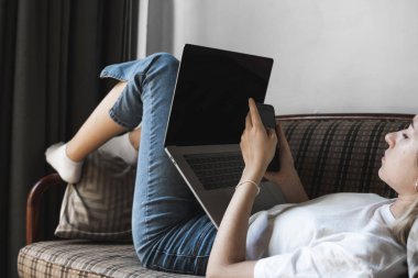 Dizüstü bilgisayarı olan bir kadın kanepede uzanıyor ve akıllı telefon kullanıyor. İnternette çalış ve çalış, serbest çalış. Defteriyle çalışan serbest çalışan bir kız elinde telefonuyla kanepede yatıyor..