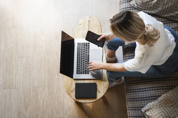 Mujer con un ordenador portátil está sentado en un sofá y utilizar un teléfono inteligente. Estudiar y trabajar en línea, freelance. Chica autónoma está trabajando con su cuaderno sentado en un sofá con un teléfono y leer en la mesa . — Foto de Stock