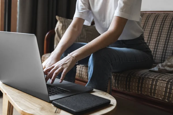 Mujer está sentado en un sofá y el uso de un ordenador portátil en la mesa de madera. Estudiar y trabajar en línea, freelance. Chica autónoma está trabajando con su cuaderno sentado en un sofá con un teléfono y leer en la mesa . — Foto de Stock