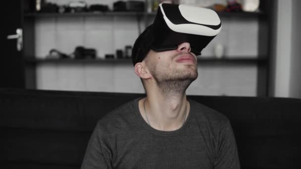 Joven barbudo con auriculares de realidad virtual en la oficina sentado en un sofá y tratando de tocar algo que ve en el mundo virtual . — Vídeo de stock