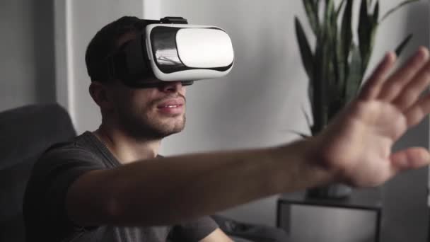 Joven barbudo con auriculares de realidad virtual en la oficina sentado en un sofá y tratando de tocar algo que ve en el mundo virtual . — Vídeos de Stock
