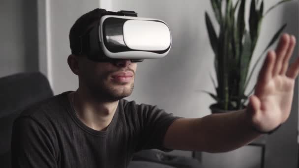 Joven barbudo con auriculares de realidad virtual en la oficina sentado en un sofá y tratando de tocar algo que ve en el mundo virtual . — Vídeos de Stock