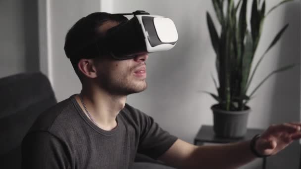 Joven barbudo con auriculares de realidad virtual en la oficina sentado en un sofá y tratando de tocar algo que ve en el mundo virtual . — Vídeo de stock