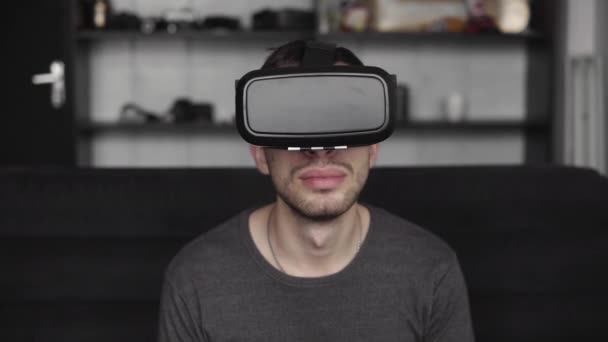 Giovane uomo barbuto che indossa cuffie di realtà virtuale in ufficio seduto su un divano. Esplorare il mondo VR e tutto ciò che ha visto in cuffia . — Video Stock