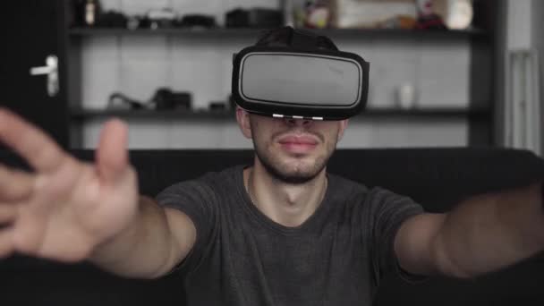 Jovem barbudo vestindo fone de ouvido de realidade virtual no escritório sentado em um sofá e tentando tocar algo que ele vê no mundo virtual . — Vídeo de Stock