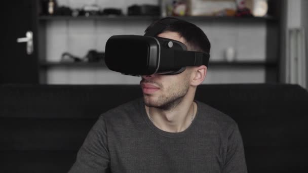 Joven barbudo con auriculares de realidad virtual en la oficina sentado en un sofá y tratando de tocar algo que ve en el mundo virtual . — Vídeo de stock