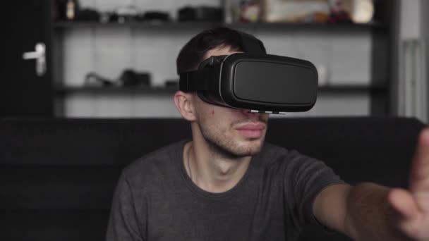 Joven barbudo con auriculares de realidad virtual en la oficina sentado en un sofá y tratando de tocar algo que ve en el mundo virtual . — Vídeo de stock