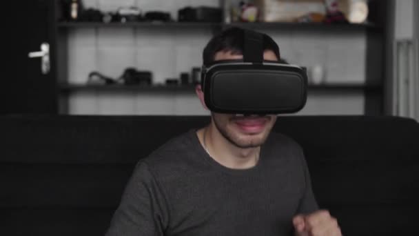 Jeune homme barbu portant un casque de réalité virtuelle au bureau assis sur un canapé et dansant . — Video