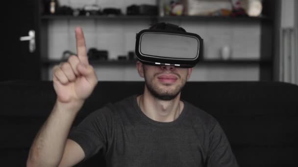 Молодий бородатий чоловік в гарнітурі віртуальної реальності сидить на дивані і намагається доторкнутися до чогось, що він бачить у віртуальному світі. Використання гарнітури VR для роботи . — стокове відео