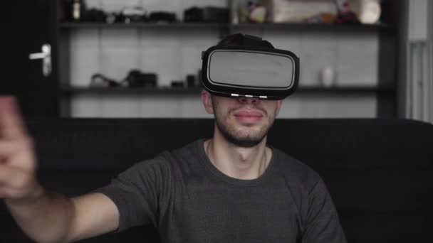 Mladý vousatý muž ve sluchátkách virtuální reality sedí na pohovce a snaží se dotknout něčeho, co vidí ve virtuálním světě. Použití Vr headsetu pro práci. — Stock video