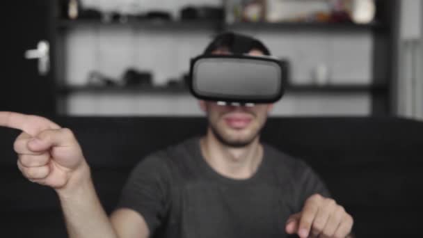 Jovem barbudo vestindo fone de ouvido de realidade virtual no escritório sentado em um sofá e tentando tocar algo que ele vê no mundo virtual. Usando fone de ouvido VR para o trabalho . — Vídeo de Stock