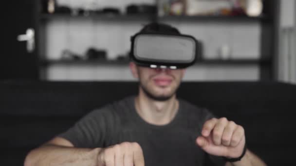 Joven barbudo con auriculares de realidad virtual en la oficina sentado en un sofá y tratando de tocar algo que ve en el mundo virtual. Uso de auriculares VR para el trabajo . — Vídeo de stock