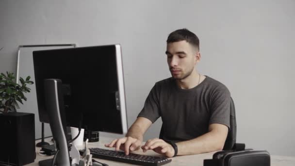 Joven estudiante de negocios o freelancer en una camisa sentada contra monitor de computadora y escribiendo un teclado en una mesa en la oficina con una expresión reflexiva. Trabajando en su portátil en la oficina . — Vídeo de stock