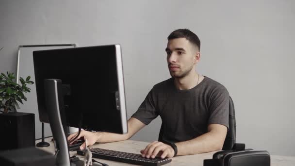 Joven estudiante de negocios o freelancer en una camisa sentada contra monitor de computadora y escribiendo un teclado en una mesa en la oficina con una expresión reflexiva. Trabajando en su portátil en la oficina . — Vídeos de Stock
