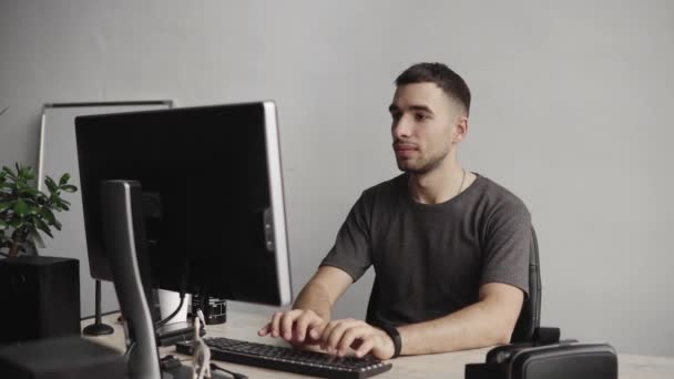 Jeune homme d'affaires étudiant ou pigiste dans une chemise assise contre le moniteur de l'ordinateur et tapant un clavier à une table dans le bureau, puis parlant par smartphone avec des partenaires d'affaires . — Video