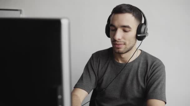 Imponerad glad leende Ung affärsman eller student i en skjorta sitter mot bildskärm i hörlurar och talar med någon via internet. — Stockvideo