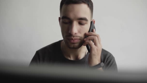 Joven estudiante de negocios o freelancer en una camisa sentada contra monitor hablando por teléfono inteligente con socios comerciales . — Vídeo de stock