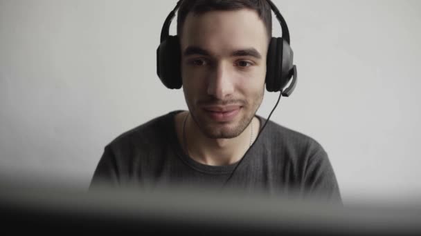 Impressionado feliz sorrindo Jovem empresário ou estudante em uma camisa sentado contra monitor de computador em fones de ouvido e falando com alguém via internet . — Vídeo de Stock