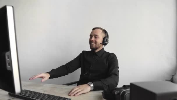 Impresionado feliz sonriente Joven hombre de negocios o estudiante en una camisa sentado contra el monitor de la computadora en los auriculares y empezar a hablar con alguien a través de Internet . — Vídeos de Stock