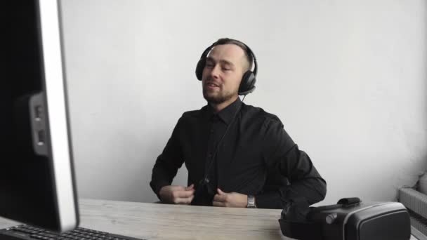 Young businessman or student in a shirt sitting against monitor of computer in office with a white background in headphones and speaking with somebody via internet with a happy face. Bussines call. — Stock Video