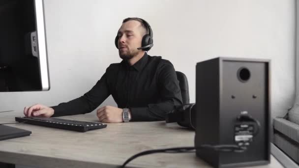 Молодой бизнесмен или студент в рубашке сидит напротив монитора компьютера в офисе с белым фоном в наушниках и разговаривает с кем-то через интернет с счастливым лицом. Звонят бизнесмены . — стоковое видео