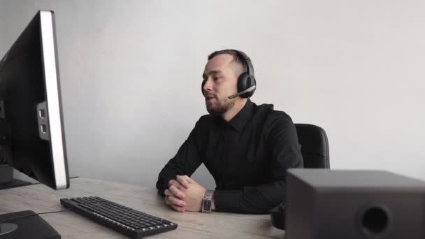Imponerad glad leende Ung affärsman eller student i en skjorta sitter mot bildskärm av datorn i hörlurar och börja tala med någon via internet. — Stockvideo