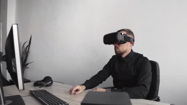 Joven barbudo sentado contra la computadora con gafas de realidad virtual tratando de tocar algo o tocar algo delante de lo que ve. Tecnologías modernas. El concepto de tecnología futura . — Vídeos de Stock