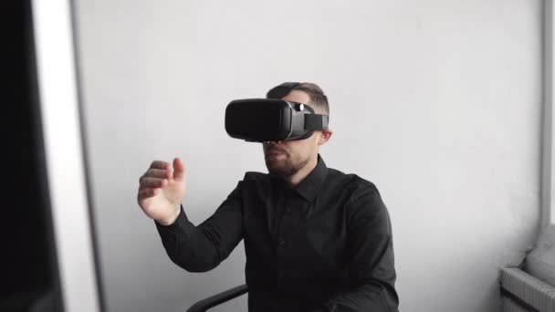 Young bearded man sitting against computer with virtual reality glasses trying to touch something or tap some thing in front of what he seeing. Modern technologies. The concept of future technology. — Stock Video